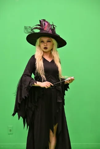 The actor in a witch costume and hat holding a wand in front of the green screen